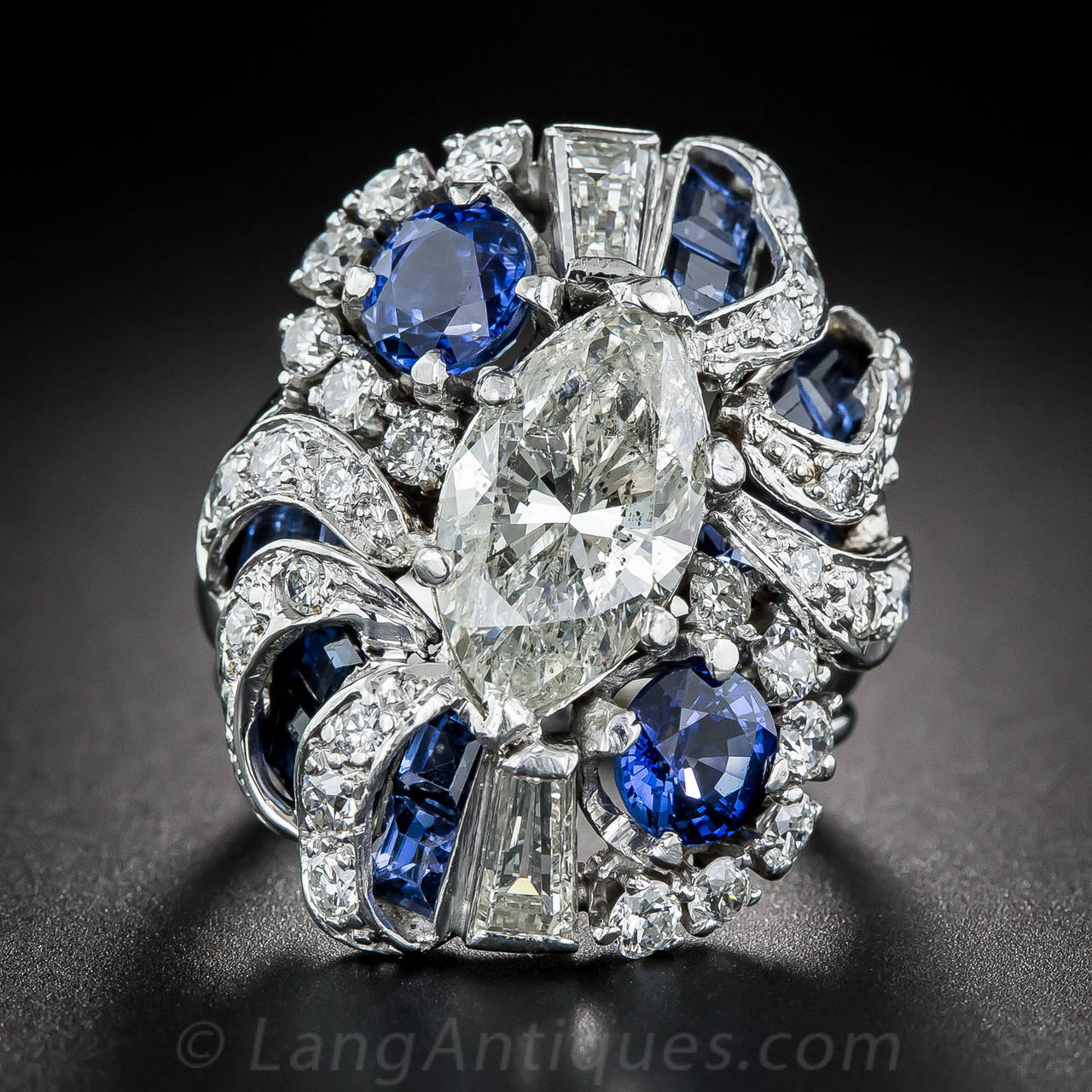 Make a sparkling statement with this 1 inch long, unique and impressive platinum, diamond and sapphire cocktail ring, starring a 3.00 carat marquise-cut diamond. The shining stone is enveloped in undulating swirls and ribbons of diamonds and is