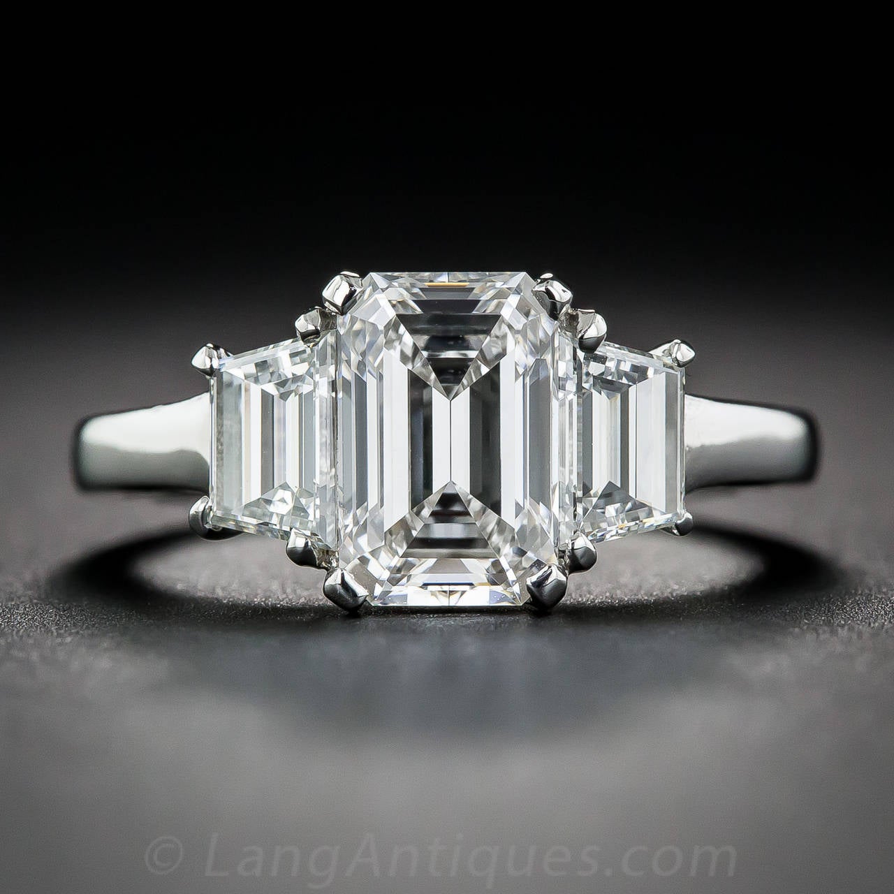 A stunning, classically modeled, emerald-cut diamond, weighing 1.61 carats, is closely embraced on each side by a matching pair trapezoid-cut diamonds (weighing an additional 1.00 carat), creating a continuous flash and sparkle across your finger,