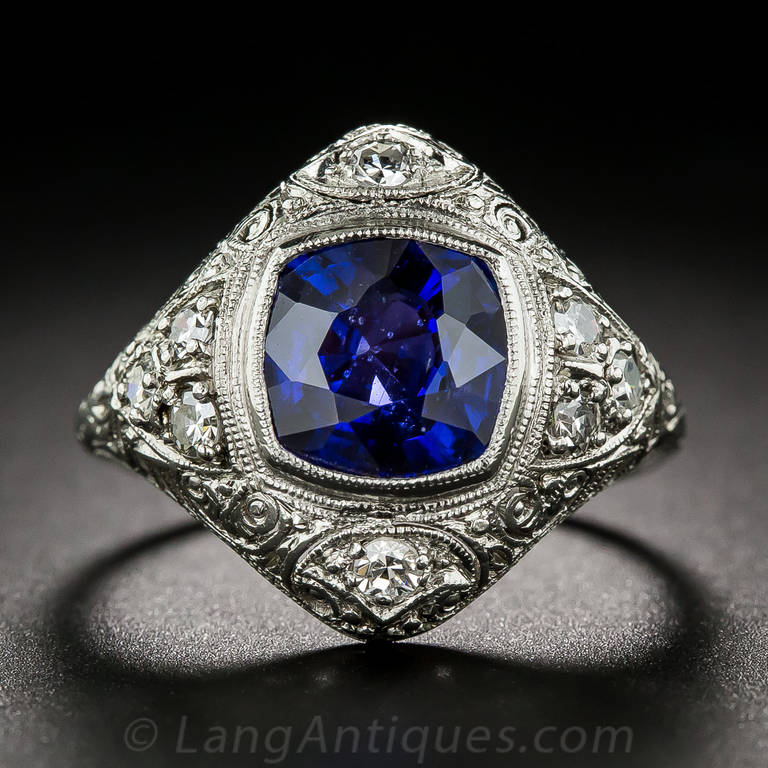 A rich royal blue cushion-cut sapphire, weighing 3.00 carats, radiates from within a finely millegrained bezel-setting, in the center of a billowy lozenge shape platinum mounting, glittering on all sides with small white single-cut diamonds. The