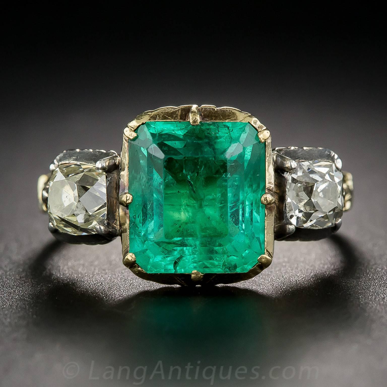 A gorgeous green Colombian emerald, weighing approximately 2.50 carats, is accompanied right and left by a pair of sparkling antique cushion-cut diamonds, dating back to the turn-of-the-twentieth-century. The emerald casts an extra glow due to the