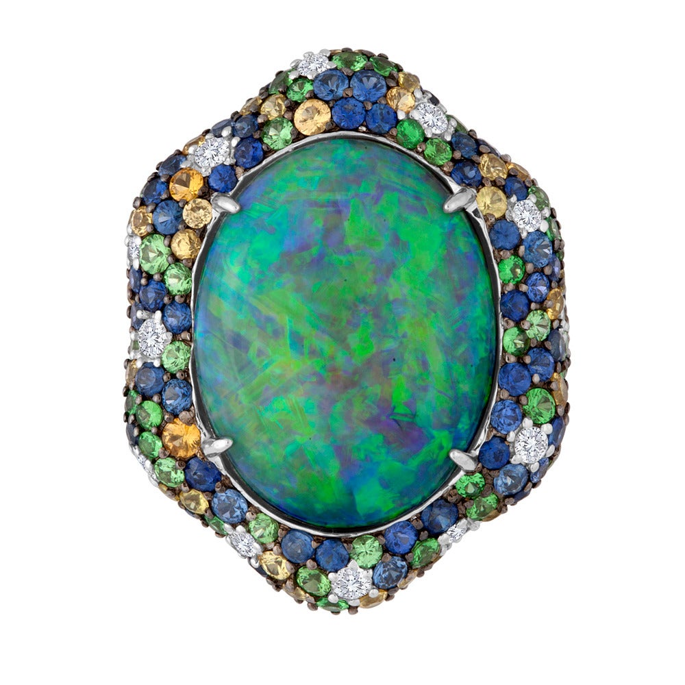 This one-of-a-kind design features an Australian black opal that presents us with deep colors of blue and green. The center stone is prong-set in an 18k white gold setting that is studded with a variety of precious gemstones that include diamonds,