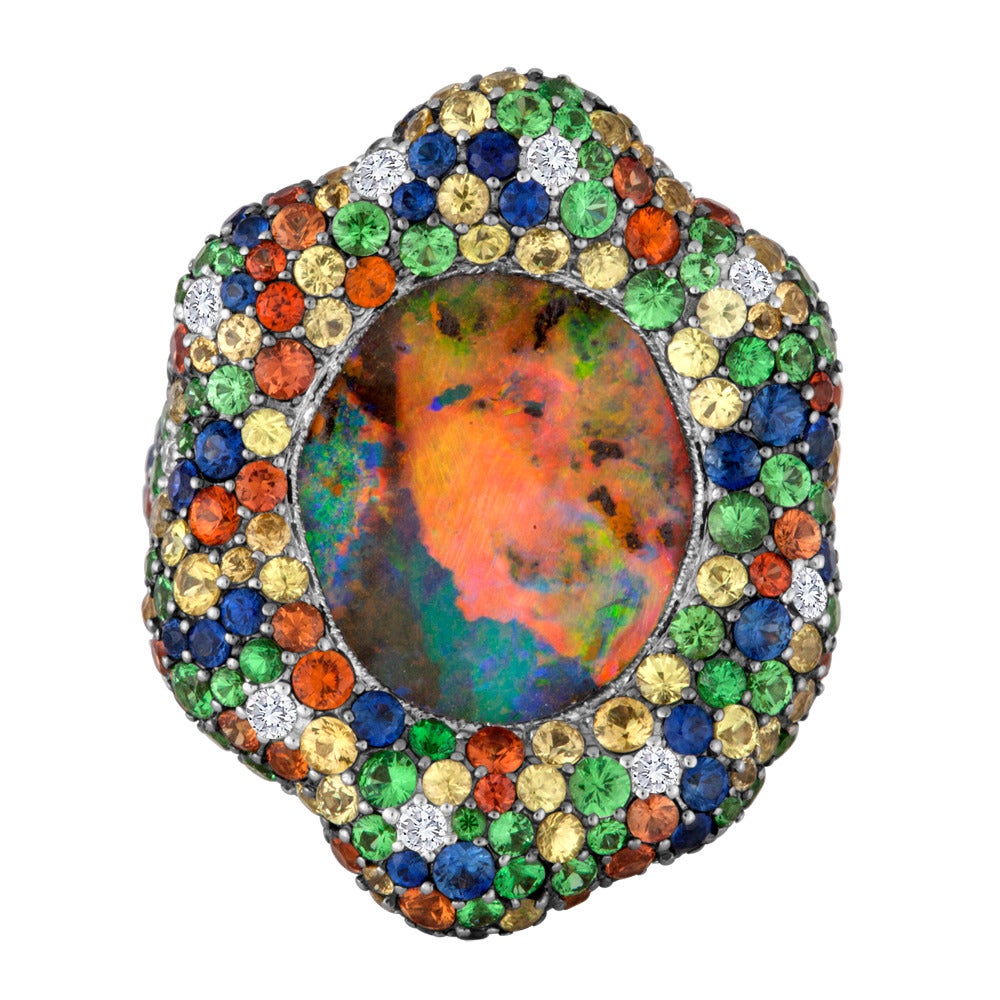 This design features an Australian black opal that presents us with a play of color that ranges from green and blue to pink and red. The center stone is prong-set in an 18k white gold setting that is studded with a variety of precious gemstones that