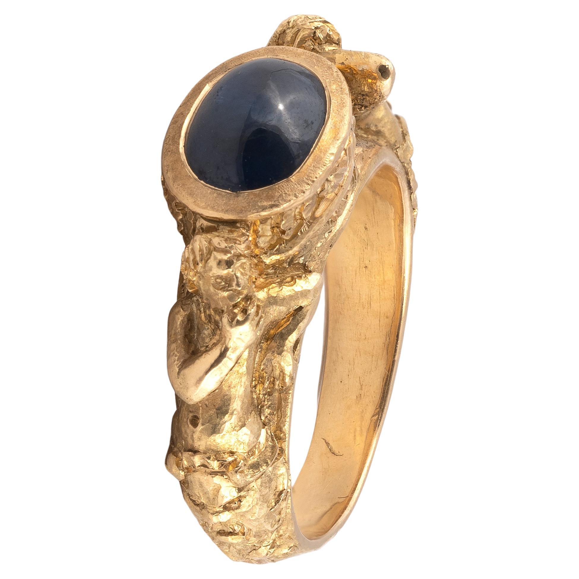 The hoop depicting a draped woman amongst foliage, her arm wrapped around a collet mounted cabochon sapphire cased.
Size 7
Weight: 13,70gr.

