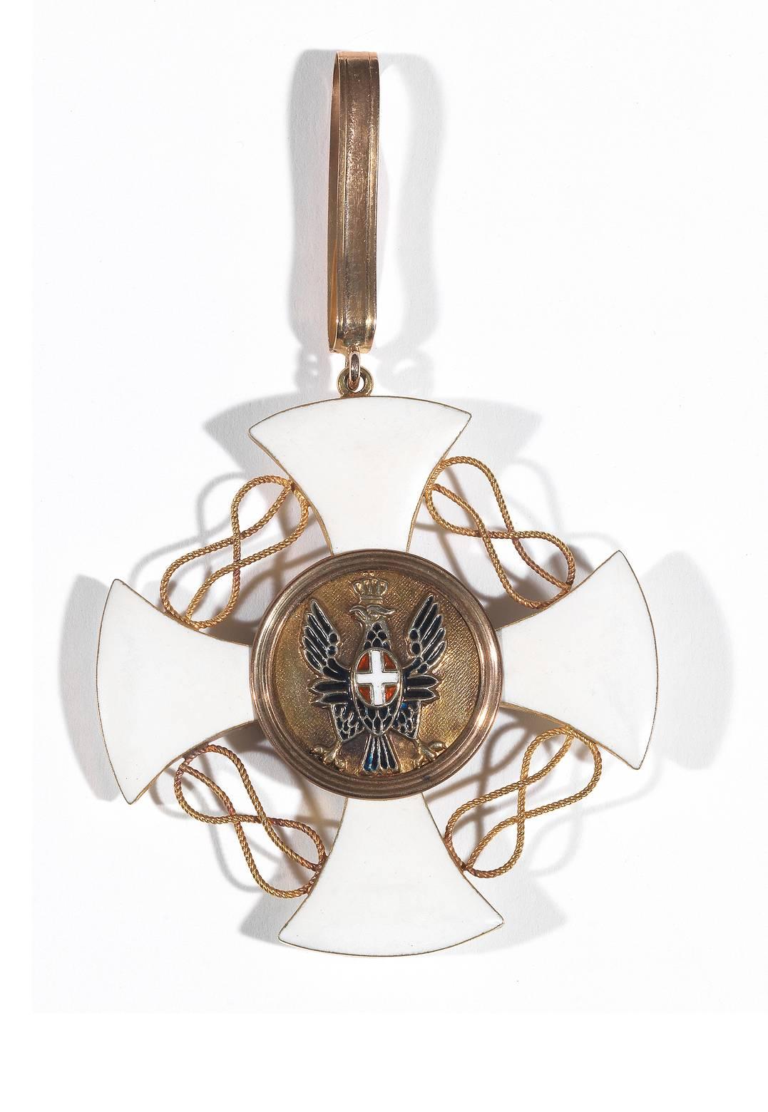 

The cross with curved edges, enamelled in white, with the so-called Savoy knots between the arms of the cross. The front central disc featured the Iron Crown of Lombardy on a blue enamel background. The back central disc with a black-enameled