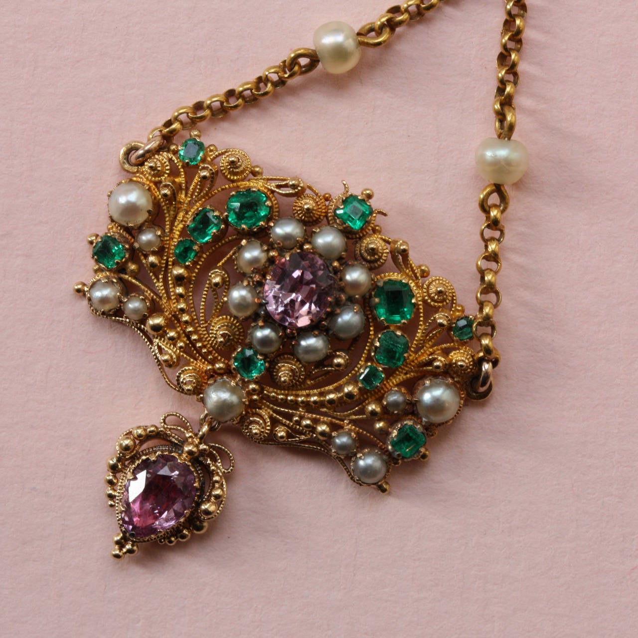 A 15 carat gold pendant decorated with fine filligree, granulations set with 12 emeralds, 19 natural pearls and 2 pink topazes, suspending from a chain.
England, circa 1820.

weight: 5.8 grams
dimensions: 5.8 x 3 cm.