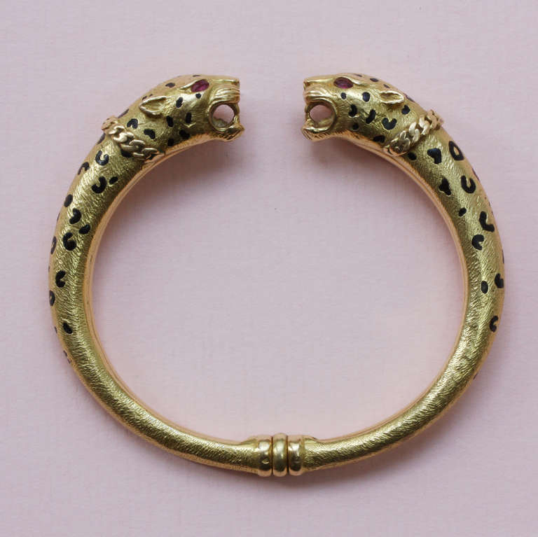 An 18 carat gold bangle with two leopard head terminals, with black enamel spots and ruby eyes, the leopards wearing gold chain collars, with French makers mark, France, circa 1970.

weight: 49 grams