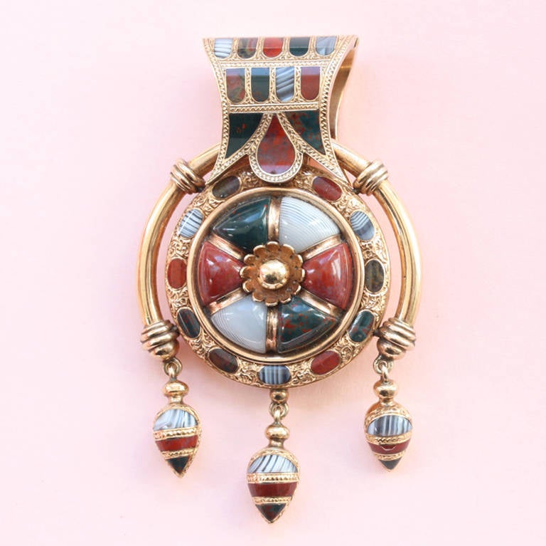 A Victorian gold and hardstone bulla with a opening locket, inlaid with agates and a gold flower at the center and three drops below, Scotland or England, circa 1870.

weight: 31 grams
dimensions: 6 x 3.5 cm.

This pendant is inspired by the