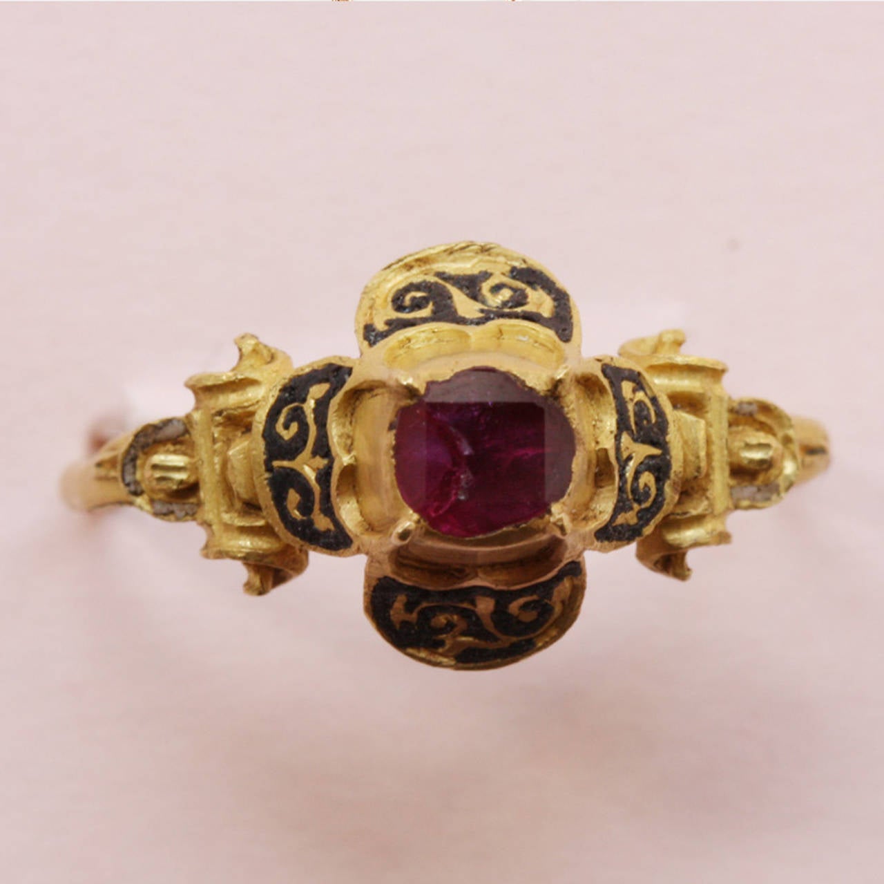 A high carat gold quatrefoil ring, the hoop merging with shoulders chased with volutes, with traces of white enamel, supporting a high quatrefoil bezel set with a table cut ruby, large black enamelled petals with foliate gold ornaments, 16th