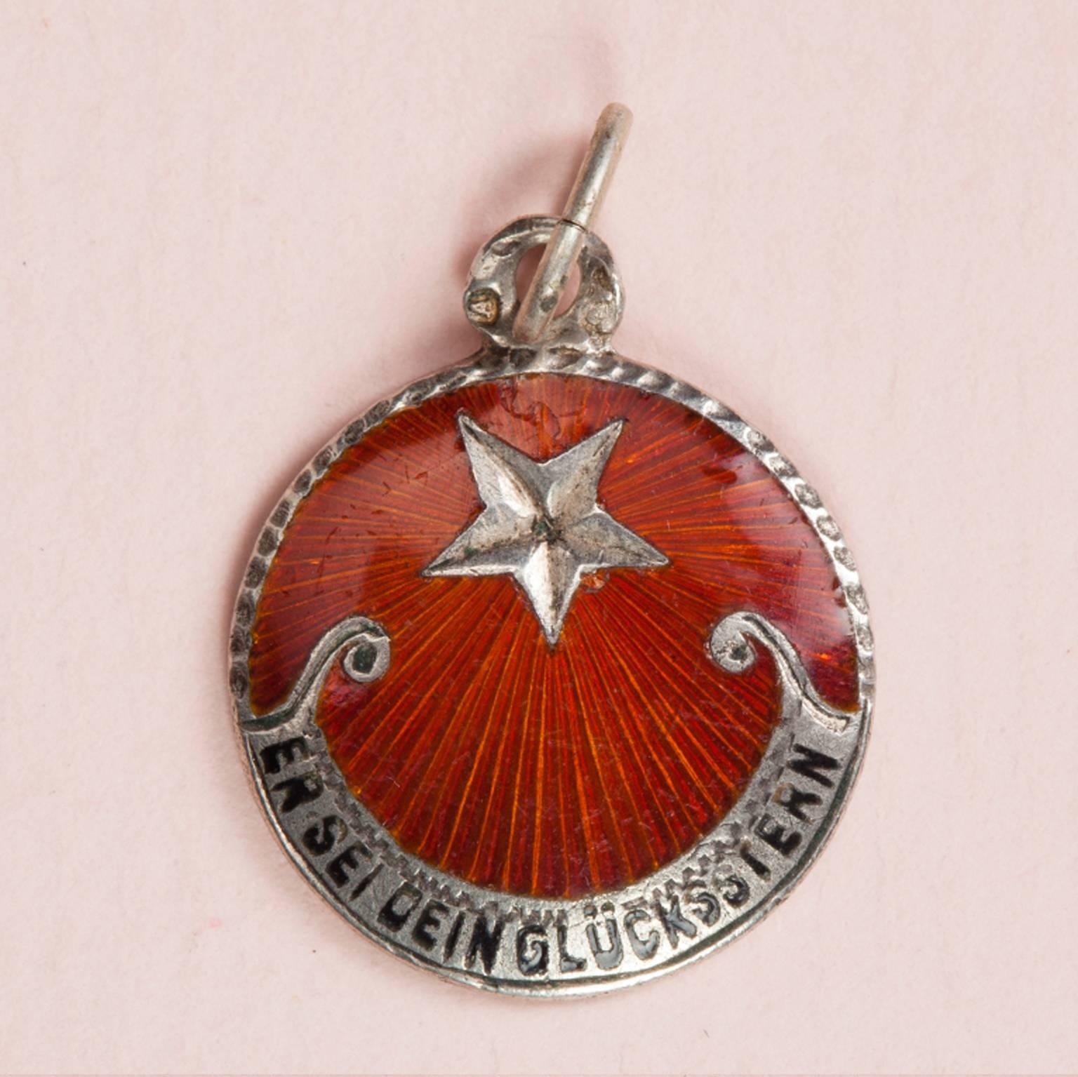 A Silver and Red Enamel Lucky Star Charm with in German 'Er Sei Dein Glücksstern' (this is your lucky star), circa 1920, Germany or Austria.

2 cm
