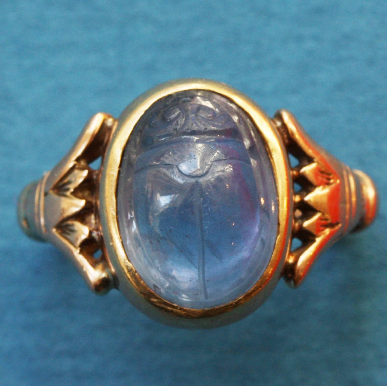 An 14 carat gold Egyptian Revival ring set with a Ceylon sapphire carved in the shape of a scarab, the inside decorated with hieroglyph inspired carvings, the shank decorated with papyrus leaves, signed: Thomas F. Brogan, New York, circa