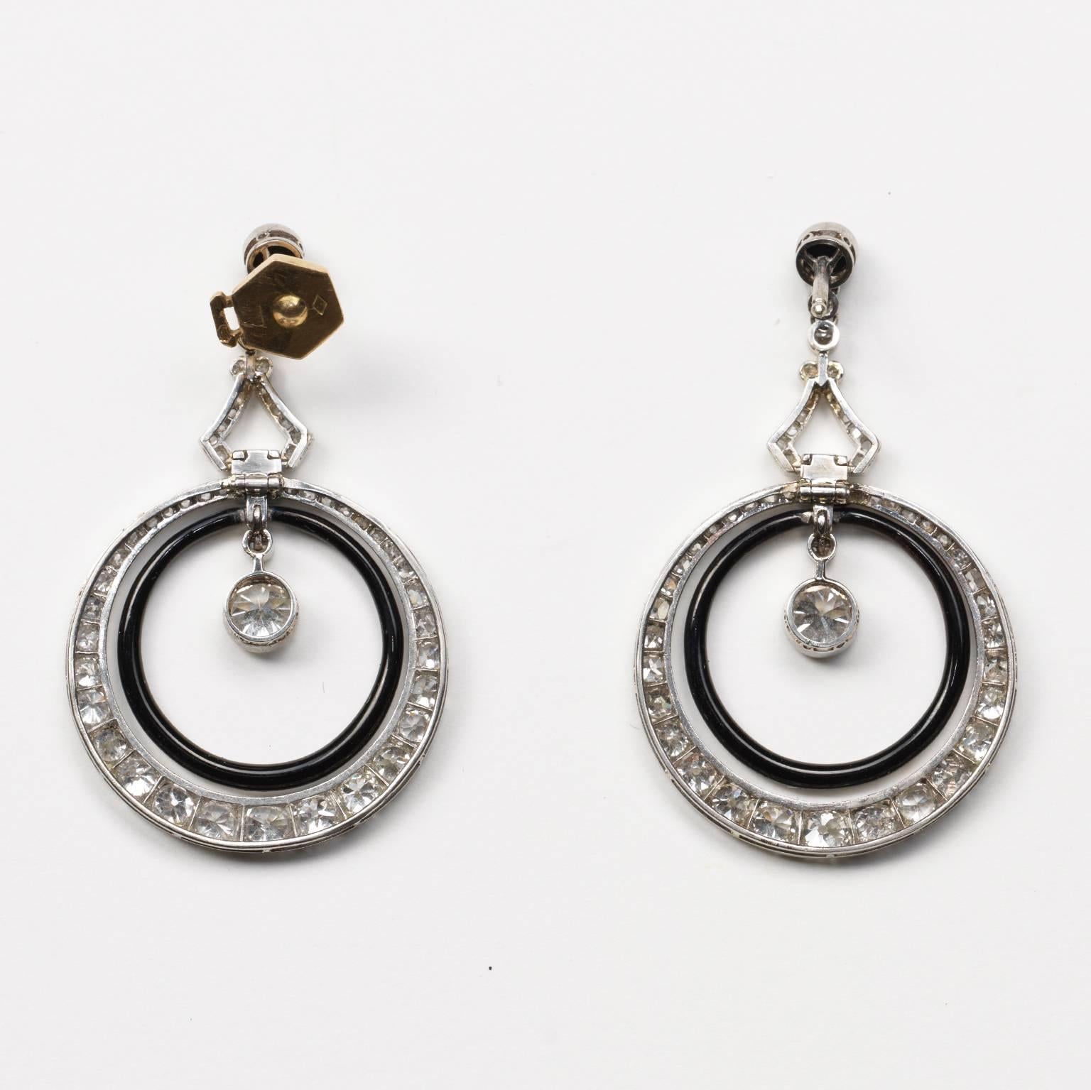 A pair of platinum and diamond (app. 3.6 carats) Art Deco earrings set with old-cut diamonds, the top is a small cabochon-cut onyx below which are two hoops connected by a triangle: the inner hoop is decorated with black enamel and the outer hoop is