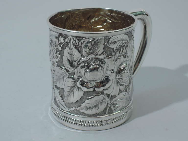 Sterling silver christening mug. Made by Gorham in Providence in 1888. Straight sides and scrolled handle. Allover floral repousse on stippled ground. Molded rim. Double beaded border at foot. Interior lightly gilt. Hallmark includes date symbol and