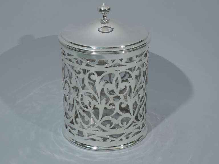 Hand-blown clear glass humidor with cased sterling silver overlay. Made by Gorham in Providence in 1911. Drum form with open scrolls, foliage, and vacant heraldic cartouche. Cover is solid silver and domed with onion finial. Cover interior has