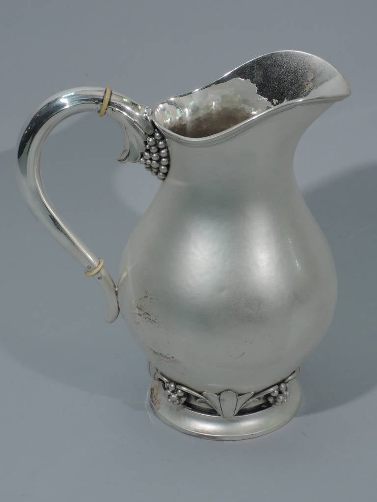 Hand-hammered sterling silver water pitcher. Made by Svend Toxvaerd in Copenhagen C 1950. Baluster body, scroll handle with grape-bunch mount, raised foot, wide lip spout, and molded rim. Base encircled with applied grape bunches alternating with