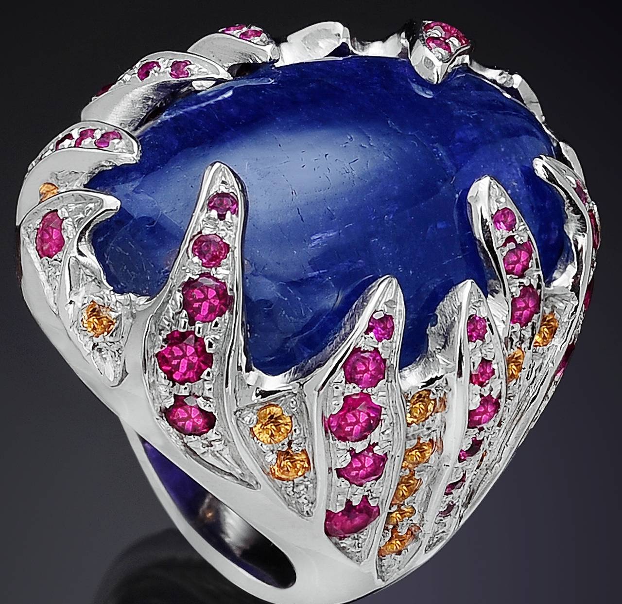 Wonderful cocktail ring inspired by fire, the 18 karat white gold encases a bold navy blue cabochon tanzanite as if protecting it from a fire, precious gemstones of rubies and yellow sapphires, representing the flames, surround it. The cabochon cut