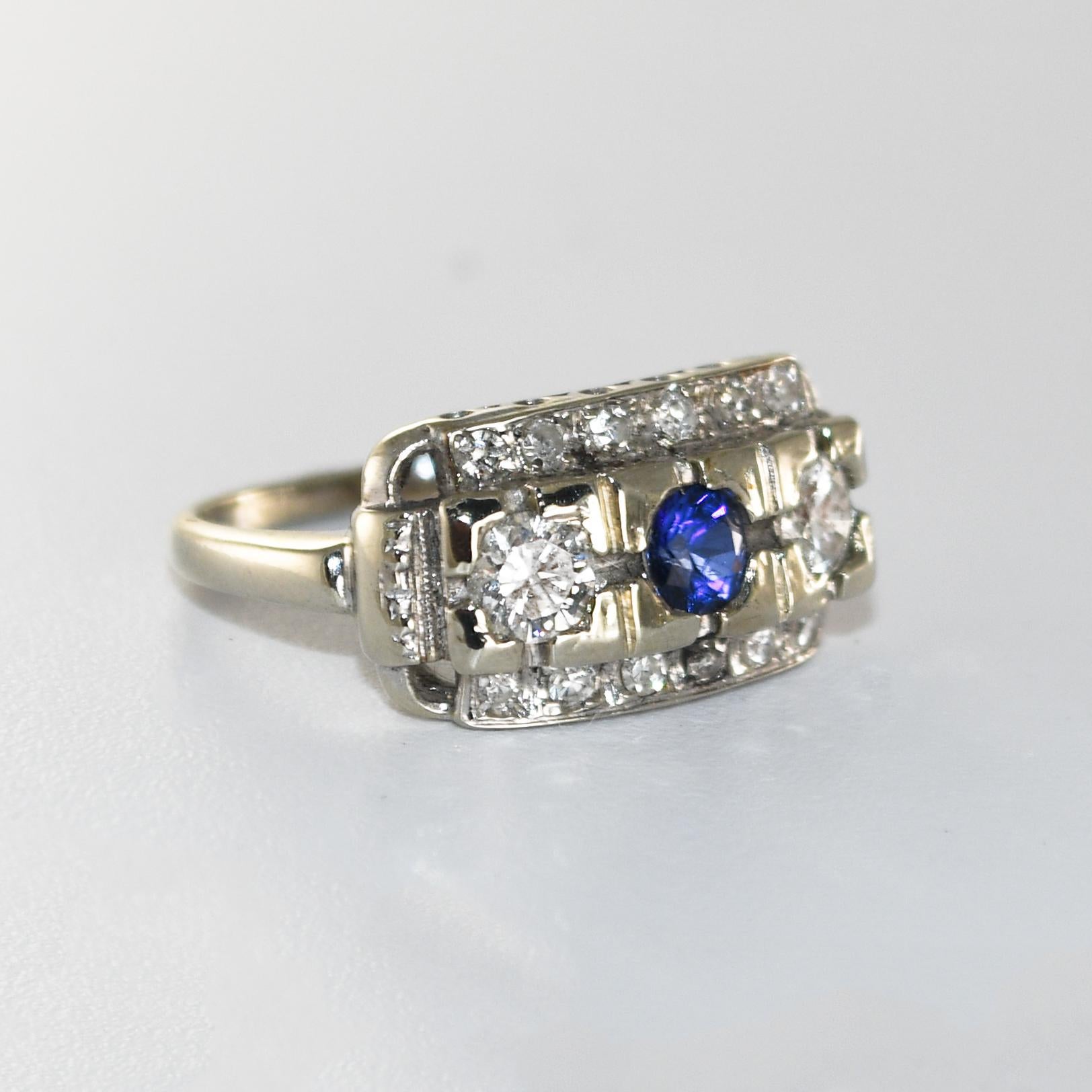 14k Weißgold Art Deco Diamant und Saphir 3 Stein Ring mit Diamant-Akzenten.
Der Mittelstein ist ein attraktiver runder blauer Saphir von 0,23 Karat. 
Auf jeder Seite des Saphirs sitzen zwei Diamanten mit einem Gesamtkaratgewicht von 0,50ct. 
Die