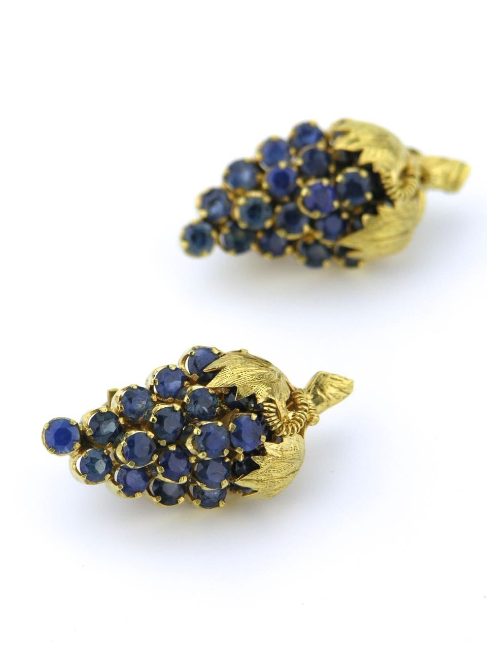 Each earring consists of a tightly set bunch of grapes motif made up of 16 round cut medium blue sapphires, capped with a pair of leaves and trailing vine - marked 14k.