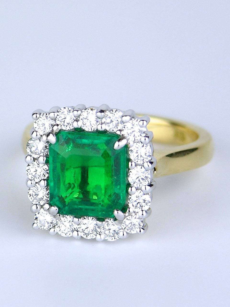 An emerald cut emerald of fine vivid green colour and medium clarity four claw set in 18ct white gold within a border of 16 round brilliant cut diamonds on an 18ct yellow gold shank 

- handmade mount 
- emerald 1=2.40cts natural, Columbian