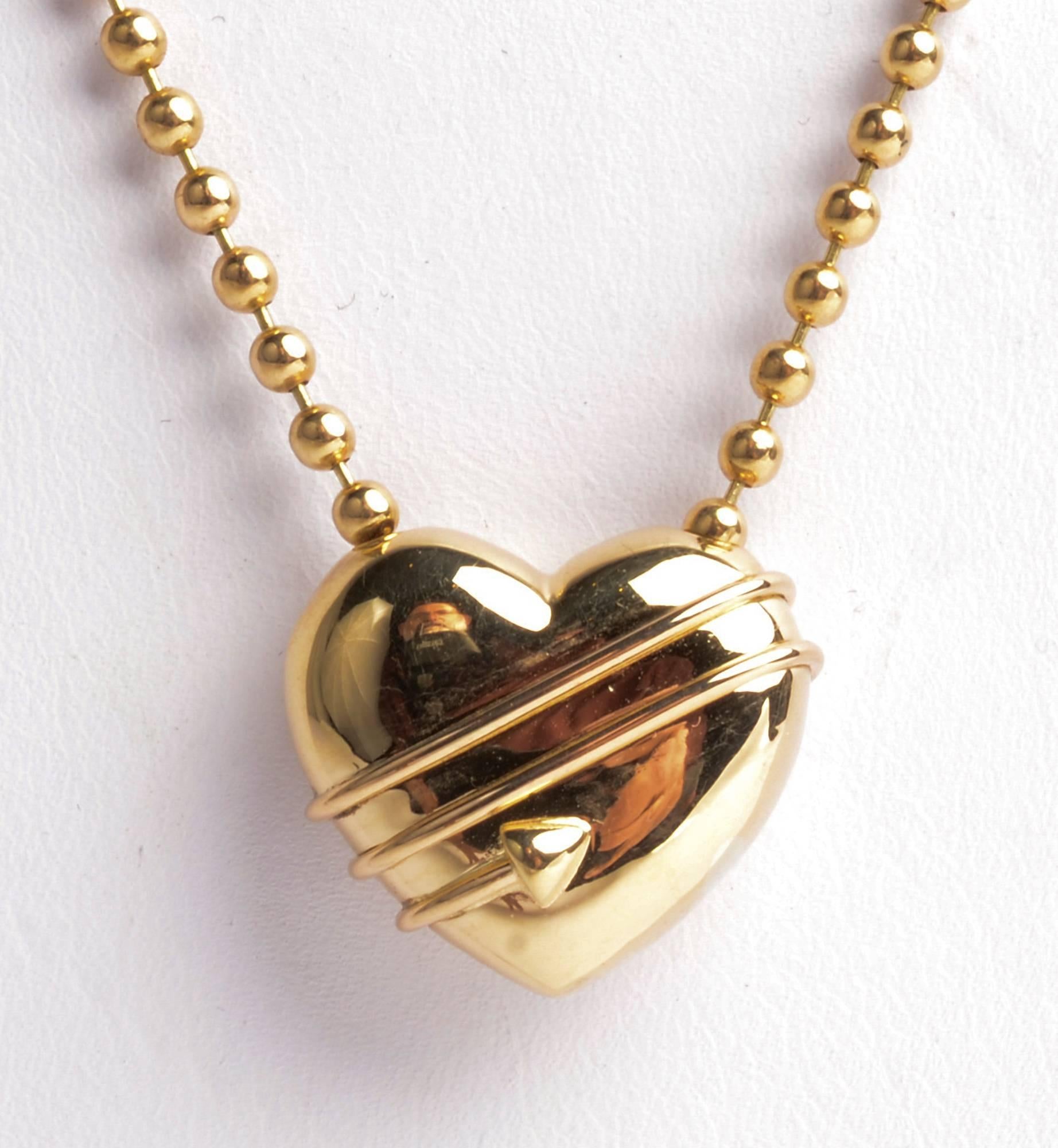 Eighteen karat gold heart pendant necklace by Tiffany. An arrow is wrapped three times around the heart. The pendant is on a Tiffany bead chain measuring 17 inches long.