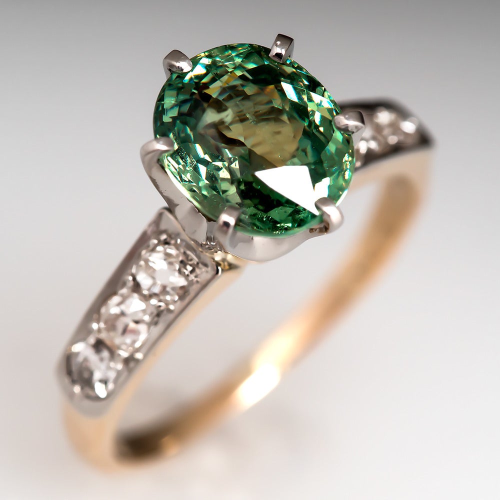 This antique demantoid garnet ring is centered with a 2.39 carat oval cut natural green garnet. Each shoulder is accented with round old European cut diamonds set into a platinum plaque. The garnet is set into a platinum crown head and the shank of