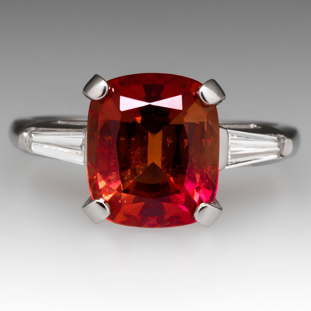 This amazing reddish-orange sapphire engagement ring is one of a kind. It is centered with a stunning GIA certified 4.73 carat natural sapphire center stone that is one of our very favorites here at EraGem. The cushion cut sapphire features a
