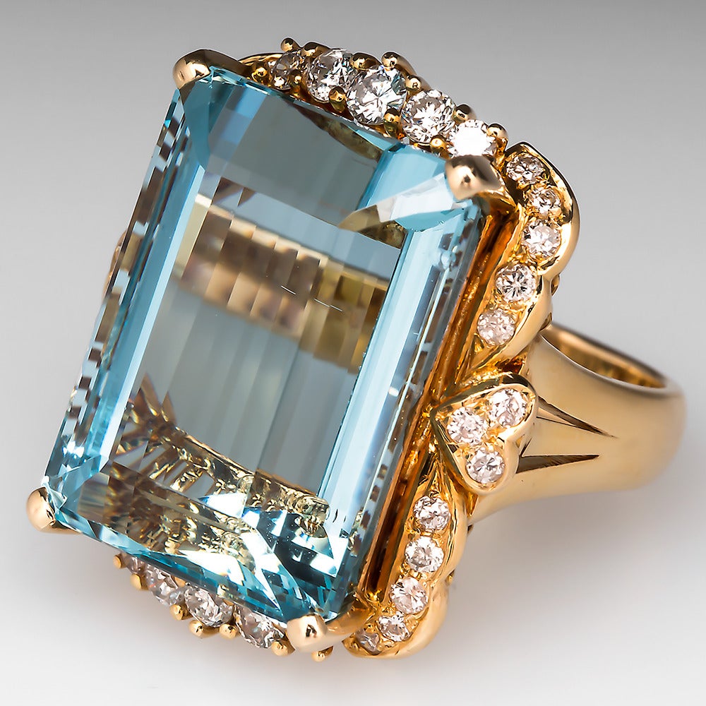 This stunning aquamarine cocktail ring features a centered emerald cut 25 carat natural aquamarine. The stone is set in a gorgeous scrolling 14k yellow gold mounting with sparkling accent diamonds. This ring is previously owned and in very good