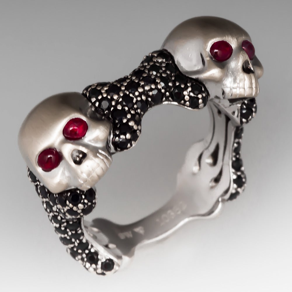 This striking Stephen Webster designer men's Skull & Bones ring is crafted of heavy solid 18k white gold. The pair of skulls have ruby eyes and the ring has pave set black sapphires. The ring is previously owned and shows some gentle wear at the