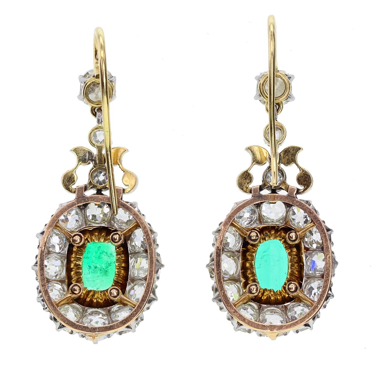 A fine and impressive pair of emerald and diamond earrings circa 1900. Each featuring a large emerald-cut emerald mounted in 18-carat gold claws, surrounded by 12 old mine-cut diamonds to form an oval cluster. Topped with a trefoil style, pierced