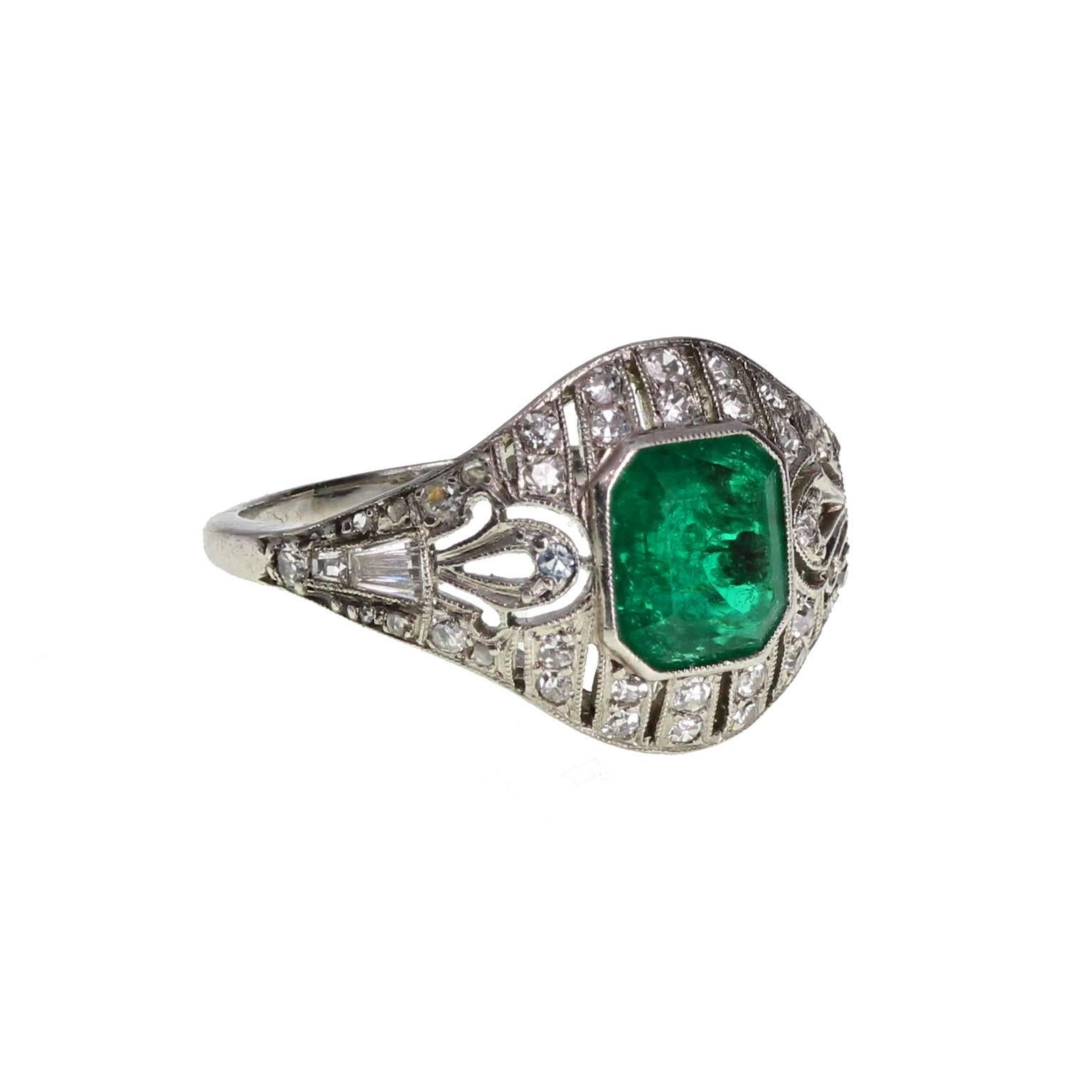 

A fine and impressive example of early 20th century style. This wonderful pierced platinum ring is decorated with single-cut and baguette-cut diamonds and detailed with mille-grain edging. A single deep-green Colombian emerald-cut emerald of