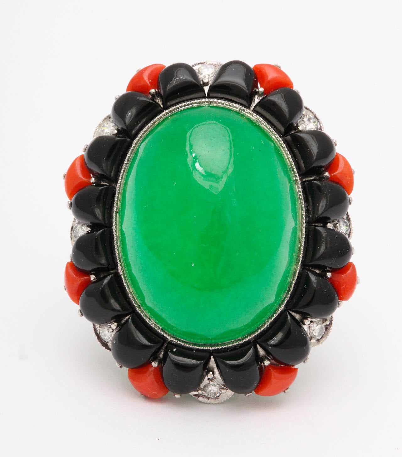 A striking ring of 18K white gold, featuring a large central cabochon of enhanced green jadeite jade, surrounded by triangular cabochons of onyx and coral, with 8 nearly colorless round brilliant cut diamonds, and 8 smaller diamonds on the shank.
