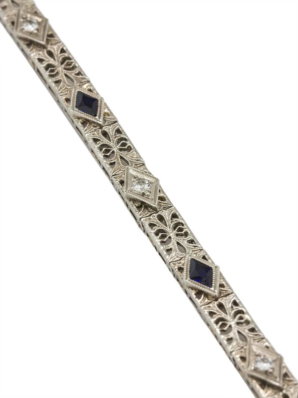 Elegant 18k white gold straight-line diamond and synthetic sapphire bracelet, with intricate filigree latticework in a romantic floral motif design. Two transitional cut round diamonds weighing approximately .15ct alternating with two synthetic