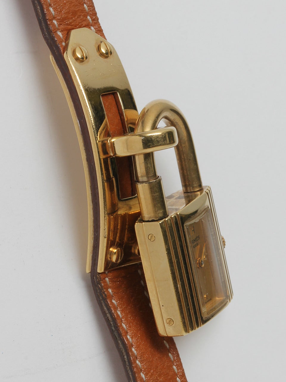 Hermes Gold Plated Kelly Lock Watch. Gold plated on stainless steel case in the shape of a lock measuring 20 x 20mm. The watch dangles from a gold plated base on tan Hermes Epsom leather strap with tang buckle. The watch features a gold color dial