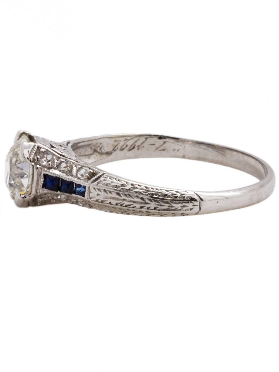 Sparkling from every direction, this stunning 0.94ct old European cut diamond, I color, SI1 clarity, sits between calibre-cut  midnight blue sapphires. This Art Deco ring is embellished by a  hand pierced gallery and hand engraved sides. A truly