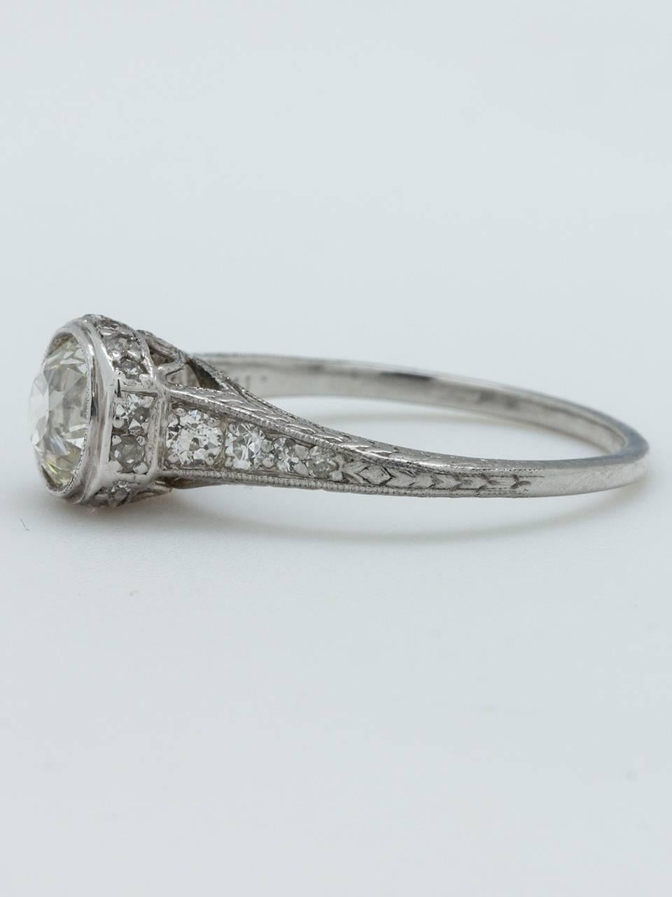 This beautifully crafted 1920s platinum engagement ring features a stunning EGL certified 0.93 carat Old European cut diamond, J-VS2. The elevated bezel setting is adorned with intricately executed wheat pattern engraving and tiny bead set side
