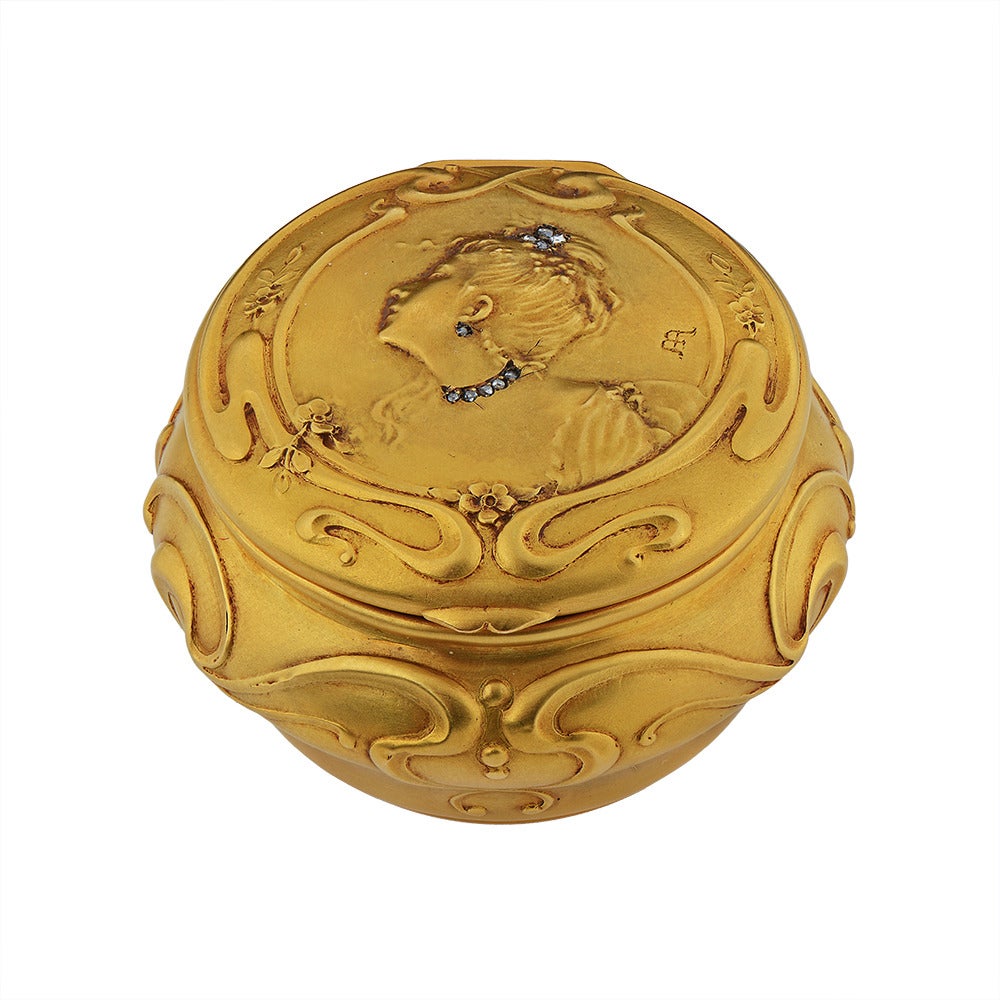 An Exquisite Art Nouveau 18ct Gold & Diamond Powder Box, French circa 1900.
The circular bombe form embossed with tendrils & scrolls, the similar hinged lid embossed with flowering foliage and an elegant “French Lady” her hair,neck & ears,