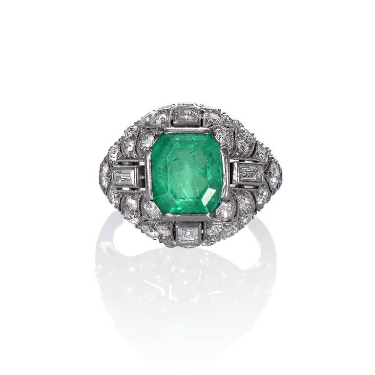 An Art Deco ring with an emerald to the centre, framed by four baguette cut diamonds and pavé-set round brilliants in a platinum mount of geometric, slightly bombé design typical of the 1920s.

Ring Size - L