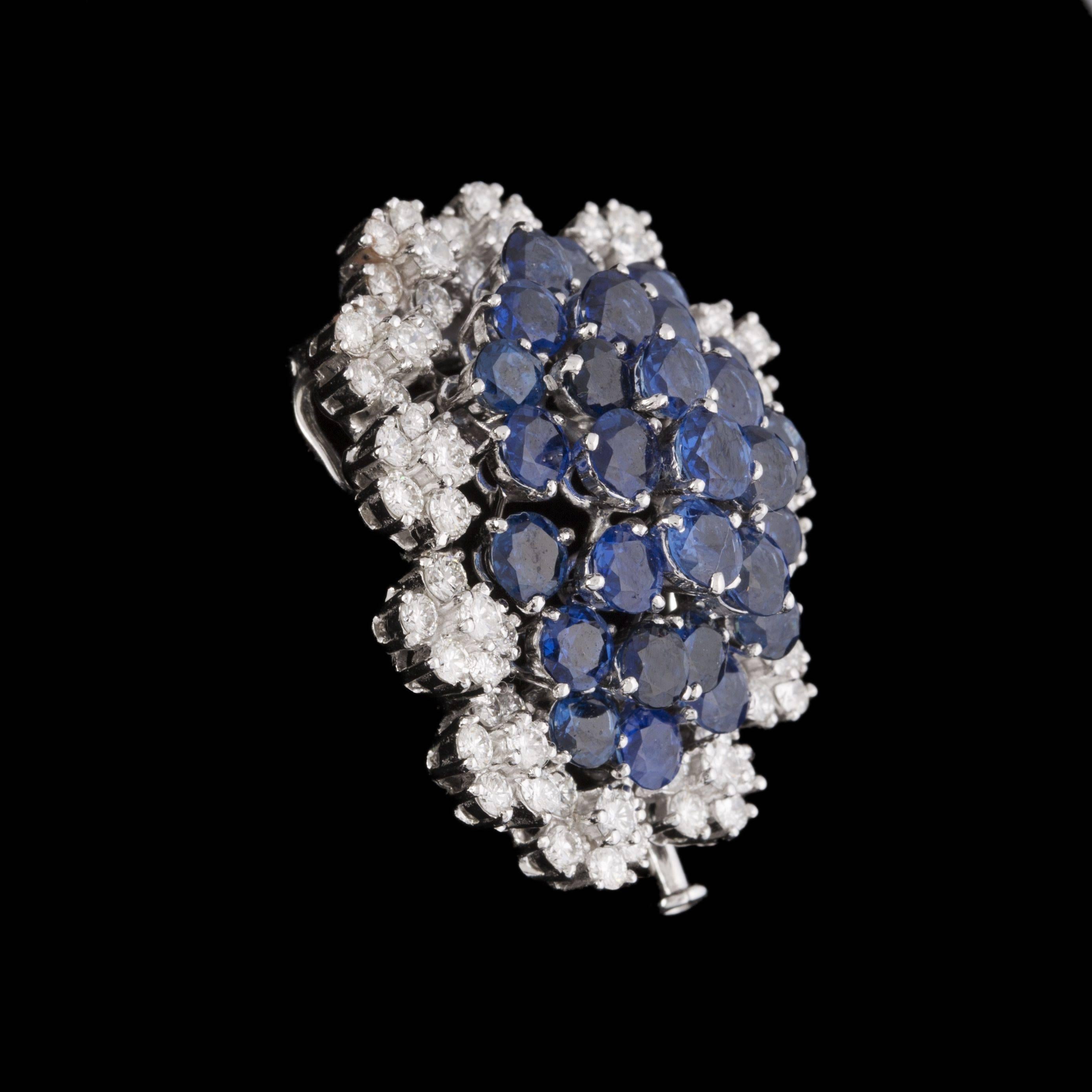 18ct (750°/oo) white gold oval and curved clip set wih round sapphires in prong setting within a surround of small brilliant-cut diamond clusters. 

This clip can be worn as a pendant. French modern work. 

Gross weight : 20.7g (0.67oz).