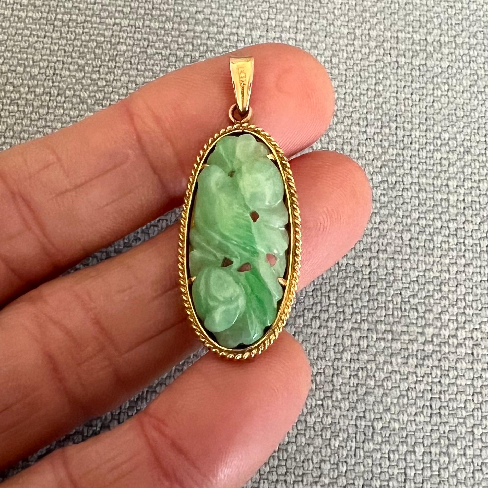A jadeite jade carved bird pendant set in a gold frame. The jadeite jade pendant is set in a 14 karat gold oval-shaped frame surrounded by a gold rope motif. The jade is beautifully carved into a floral decor with a bird resting on a branch - the