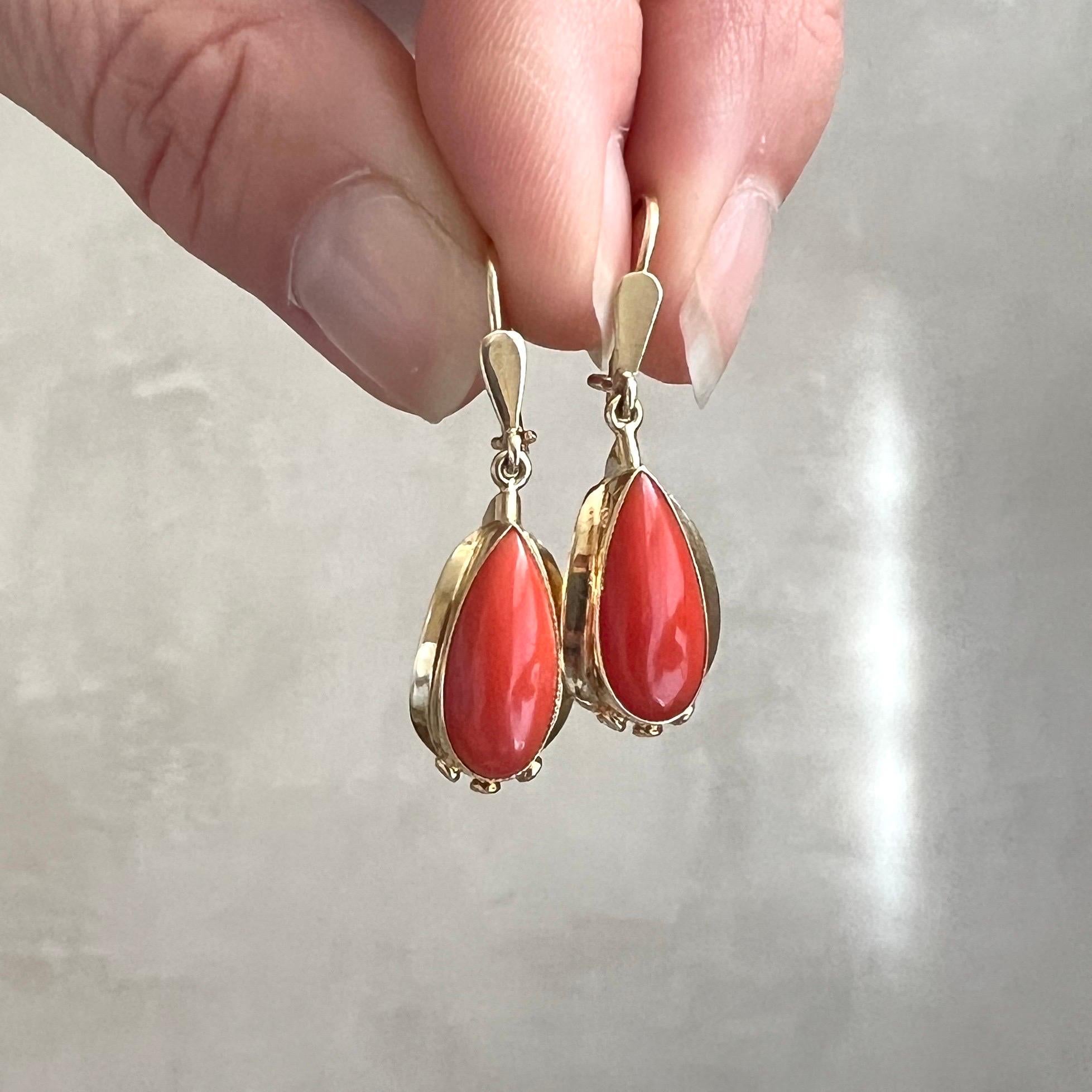 Art Deco 14 karat gold coral dangle earrings. These earrings are oval-shaped and have a smooth cabochon cut red coral mounted in 14 karat yellow gold bezel setting. At the bottom of the earrings there are three symmetrical open figures, which looks