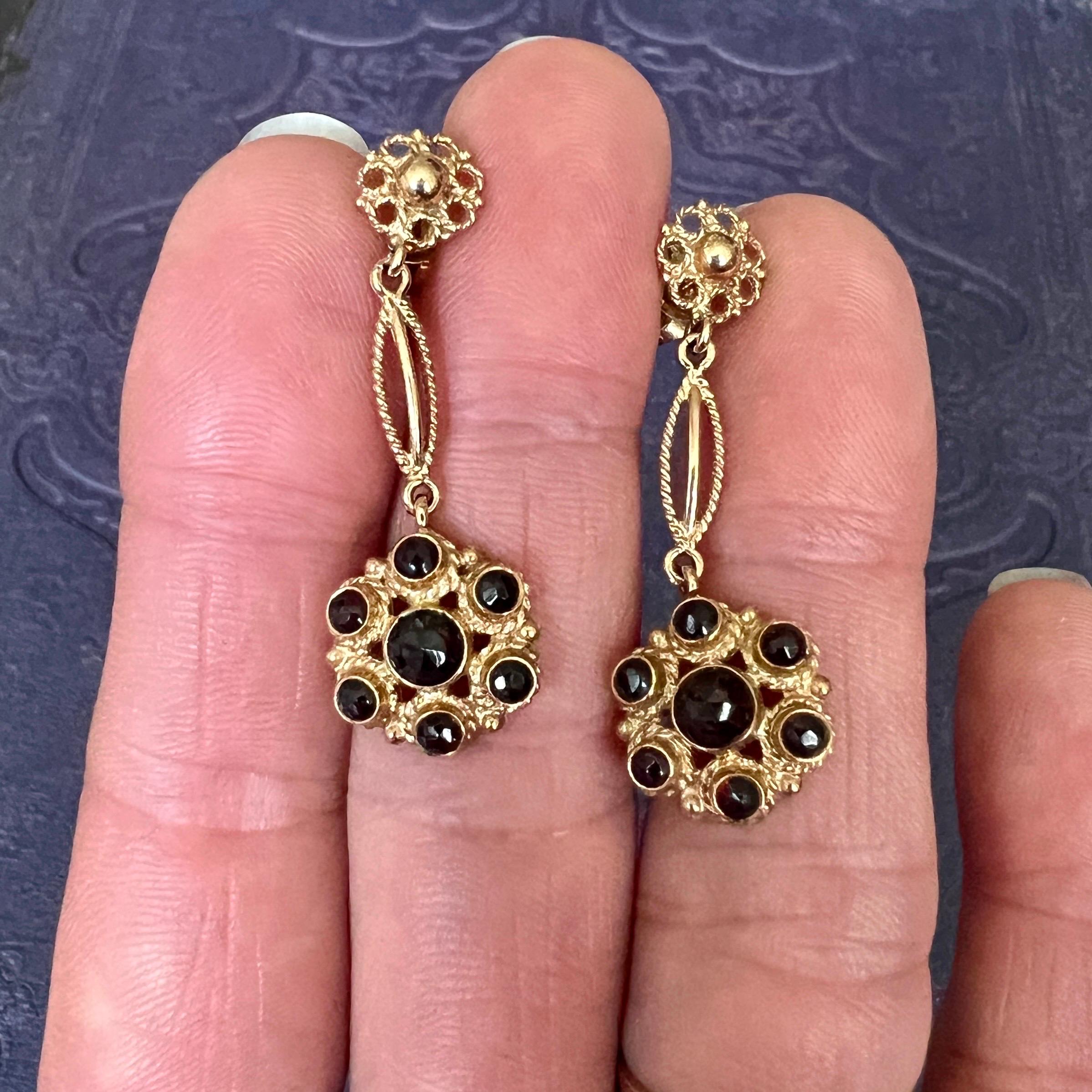 Pair of 14 karat yellow gold dangle earrings set with six faceted red garnets. The garnet stones are clustered around the center stone and bezel set. They are adorned with rope motif and granulation on the sides. The earpieces are also made of rope