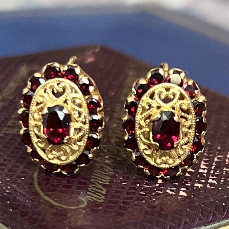 These oval-shaped drop earrings are set with a cluster of beautiful garnet stones. The center garnet is surrounded by a tangle of golden connected curls and a rim on the outer side of neatly serried garnets which are set with prongs. The garnet