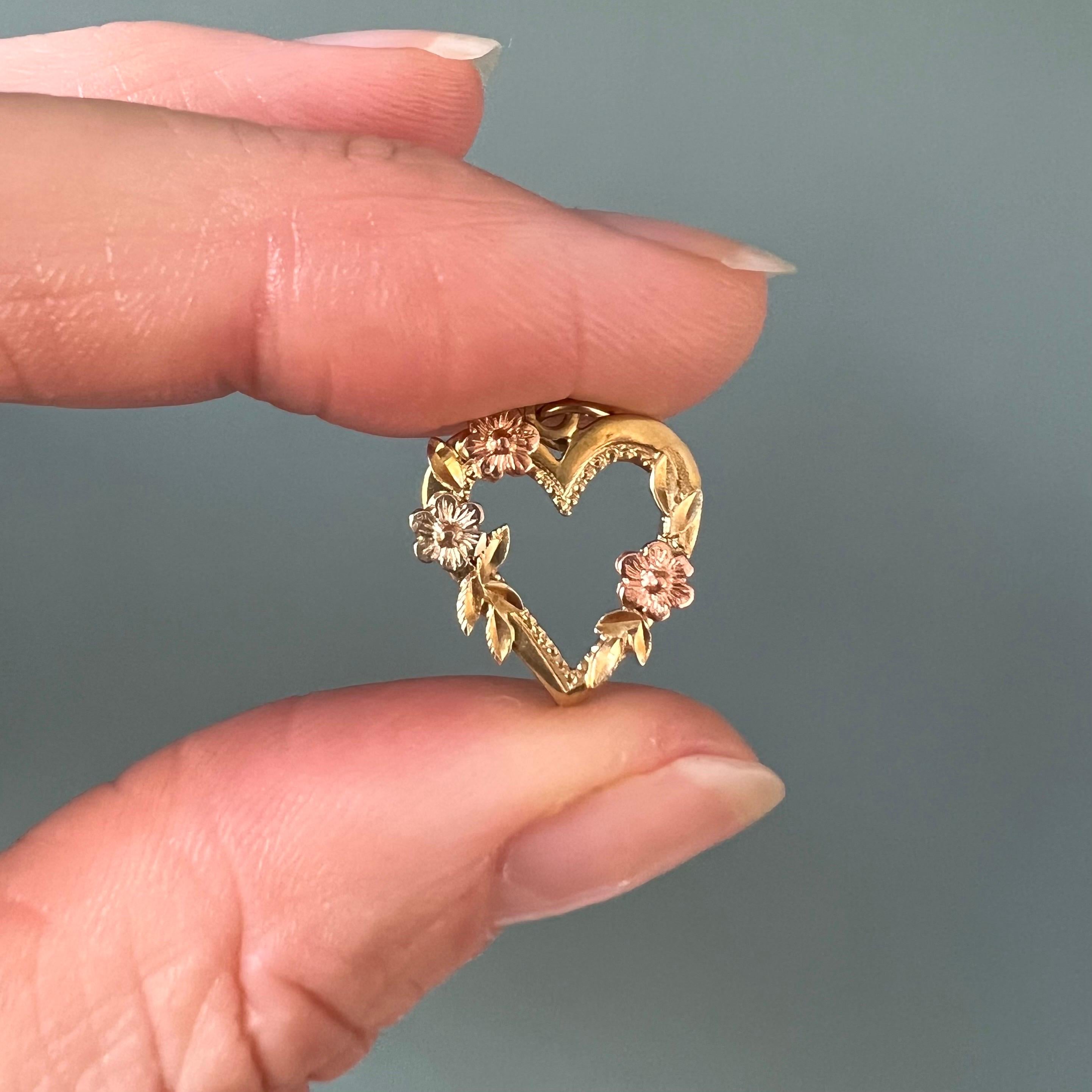 A vintage 14 karat yellow gold heart love charm pendant created with gold and rose gold flowers. The French heart is beautifully detailed with three flowers and some leaves on the border of this pretty heart. The reverse of the charm has a gold