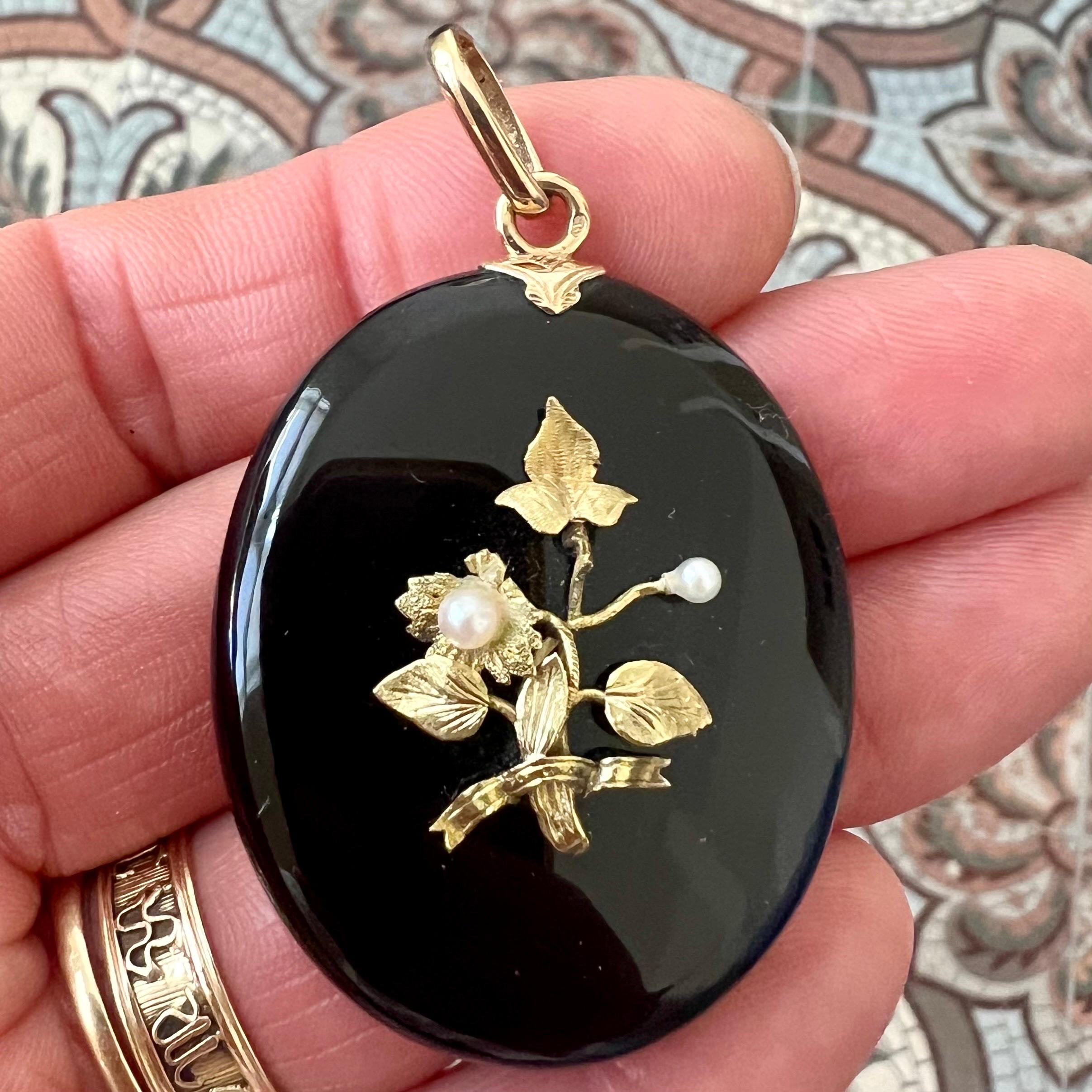 Questo bellissimo ciondolo antico a forma di medaglione ovale è finemente realizzato in onice e impreziosito da un'applicazione floreale in oro con due perle coltivate. Sul retro, il pezzo si apre per rivelare uno scomparto per le foto, che contiene