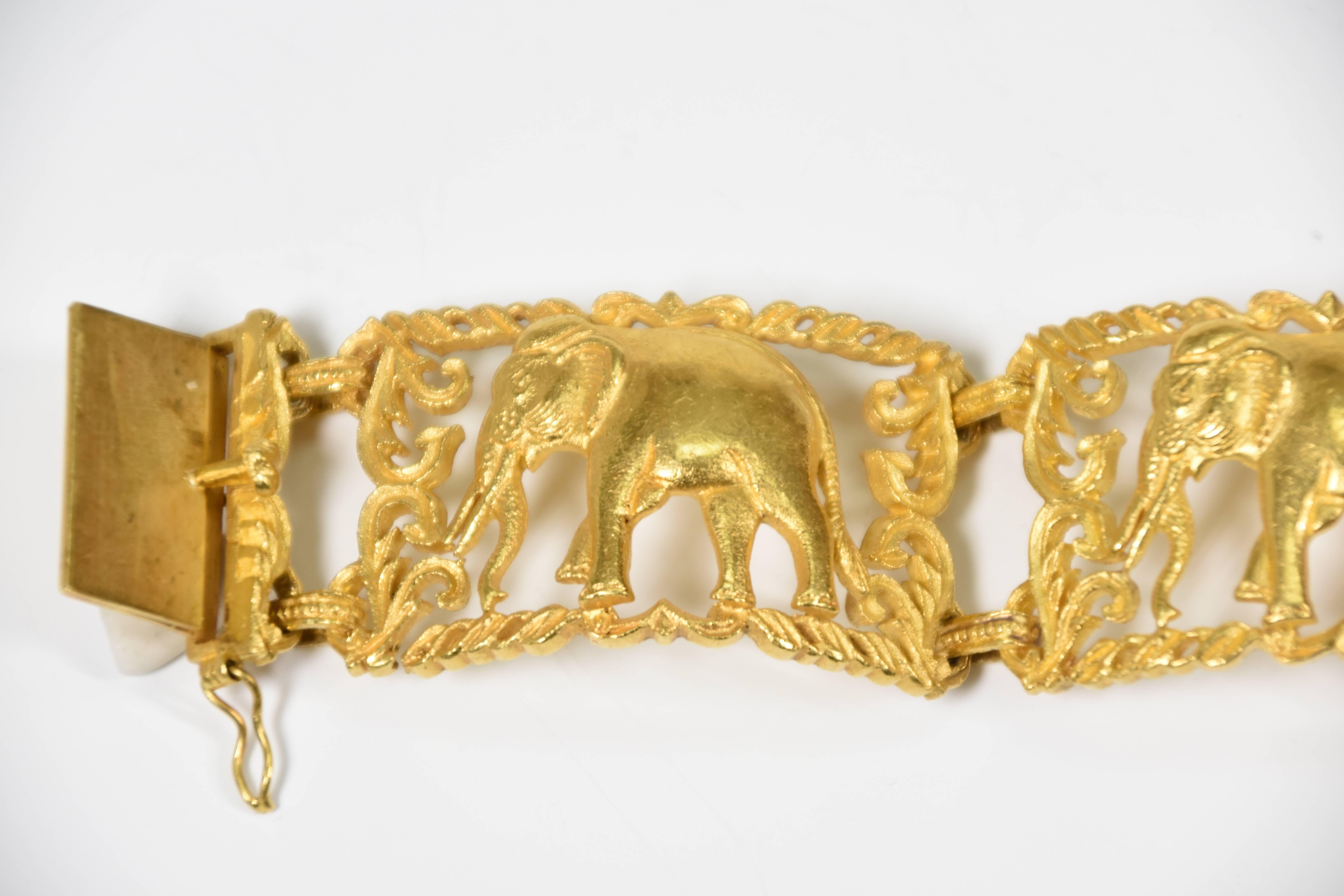 In the style of Cartier 23ct yellow gold cuff-style bracelet  featuring three individually articulated curved panels centred by an elephant motif surrounded by a scrolled and swagged border.  The bracelet measures 16.5cm in length and is fastened