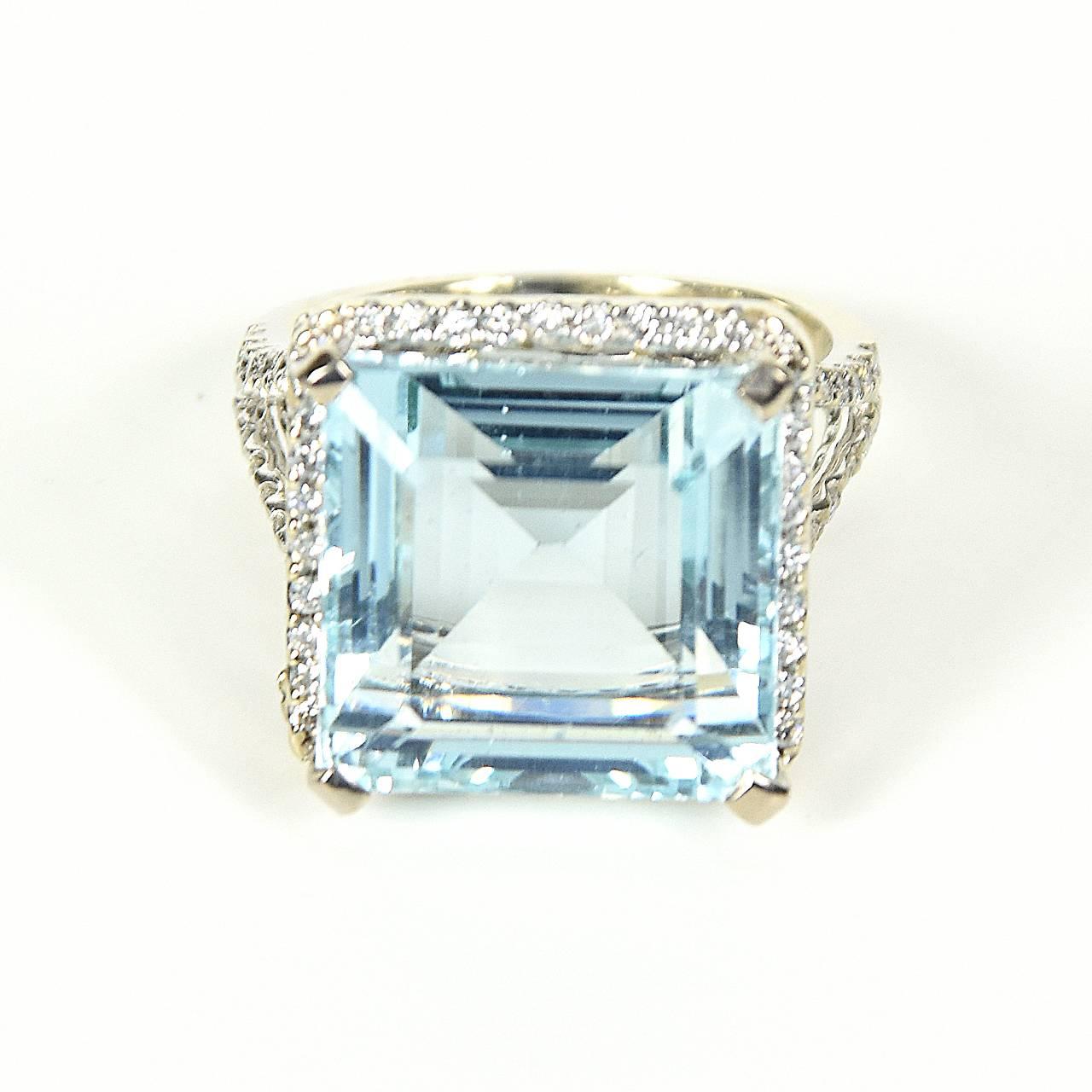 White gold dress ring featuring large square step cut light blue coloured Aquamarine 11.80 ct, secured by four claws and surrounded by a white gold railed rim containing forty small brilliant cut diamonds that are shared claw set to the perimeter.