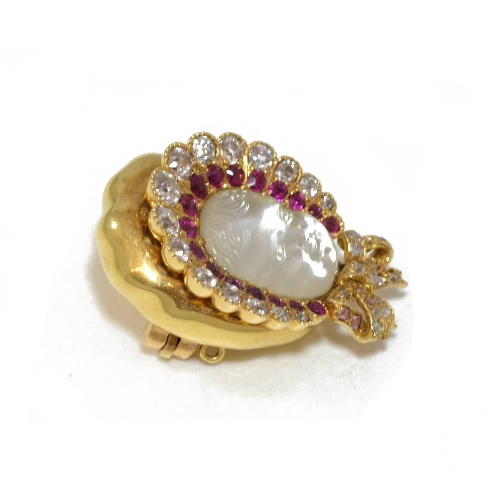A brooch in the form of a carved moonstone baby’s face, wearing a ruby and diamond set bonnet with diamond set bowed ribbon under the chin. Mounted in 18ct yellow gold. American, circa 1890.