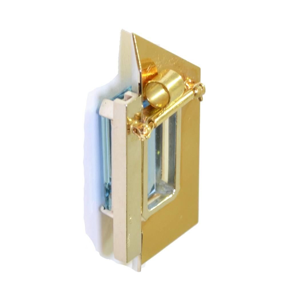 A stylised abstract brooch set with a central emerald cut aquamarine on a folded gold parallelogram with a vertical bar of 12 brilliant cut diamonds. With a detachable fitting which allows the brooch to be worn as a pendant. 14ct yellow gold.