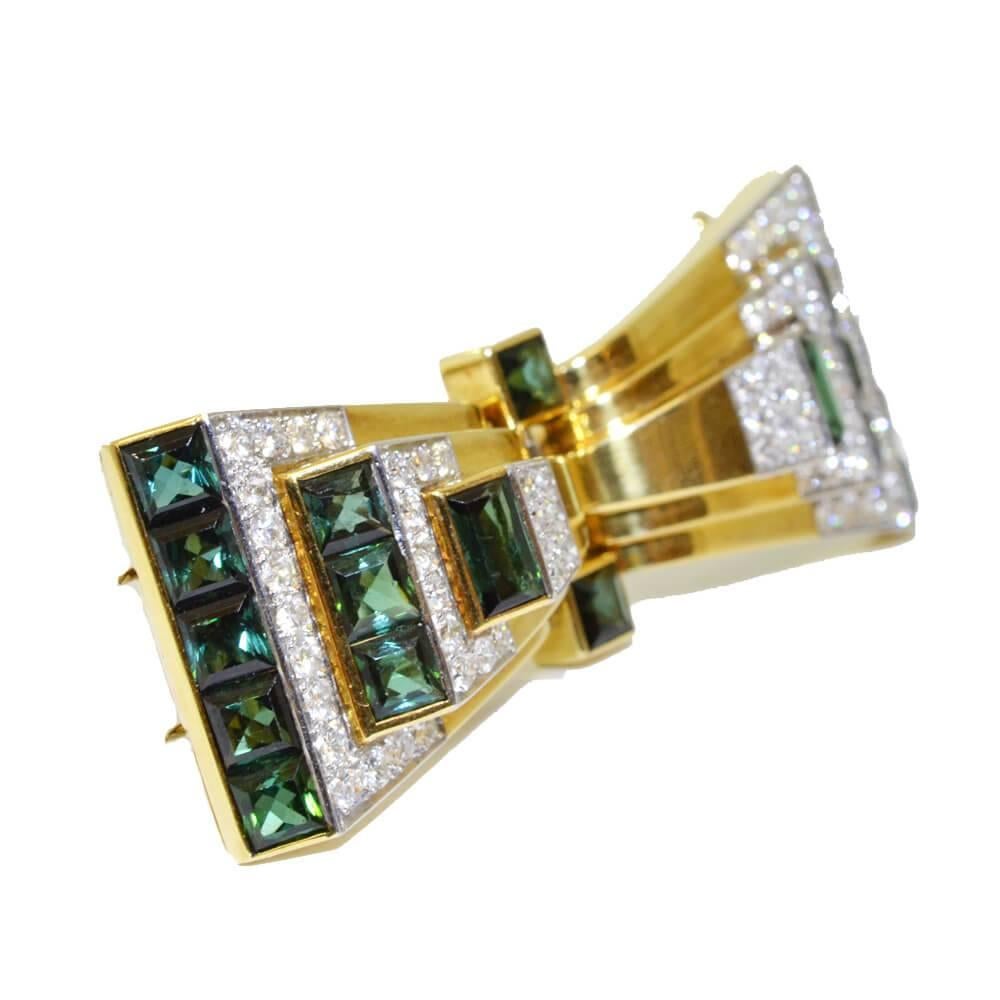 A brooch in the form of a stylised stepped bow, that separates off its frame into a pair of dress clips. Set with square cut green tourmalines to the ends of each section and brilliant cut diamonds on the front. 18ct yellow gold and platinum, by
