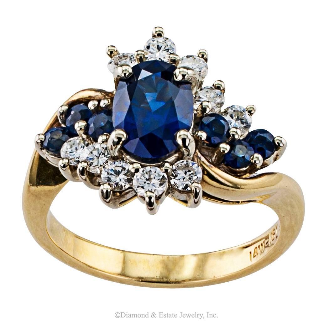 Sapphire Diamond Cluster Two Tone Gold Ring

1970s sapphire and diamond gold ring.  Centering upon an oval blue sapphire weighing approximately 1.20 caratars between shoulders alternating with six additional round-cut sapphires together weighing