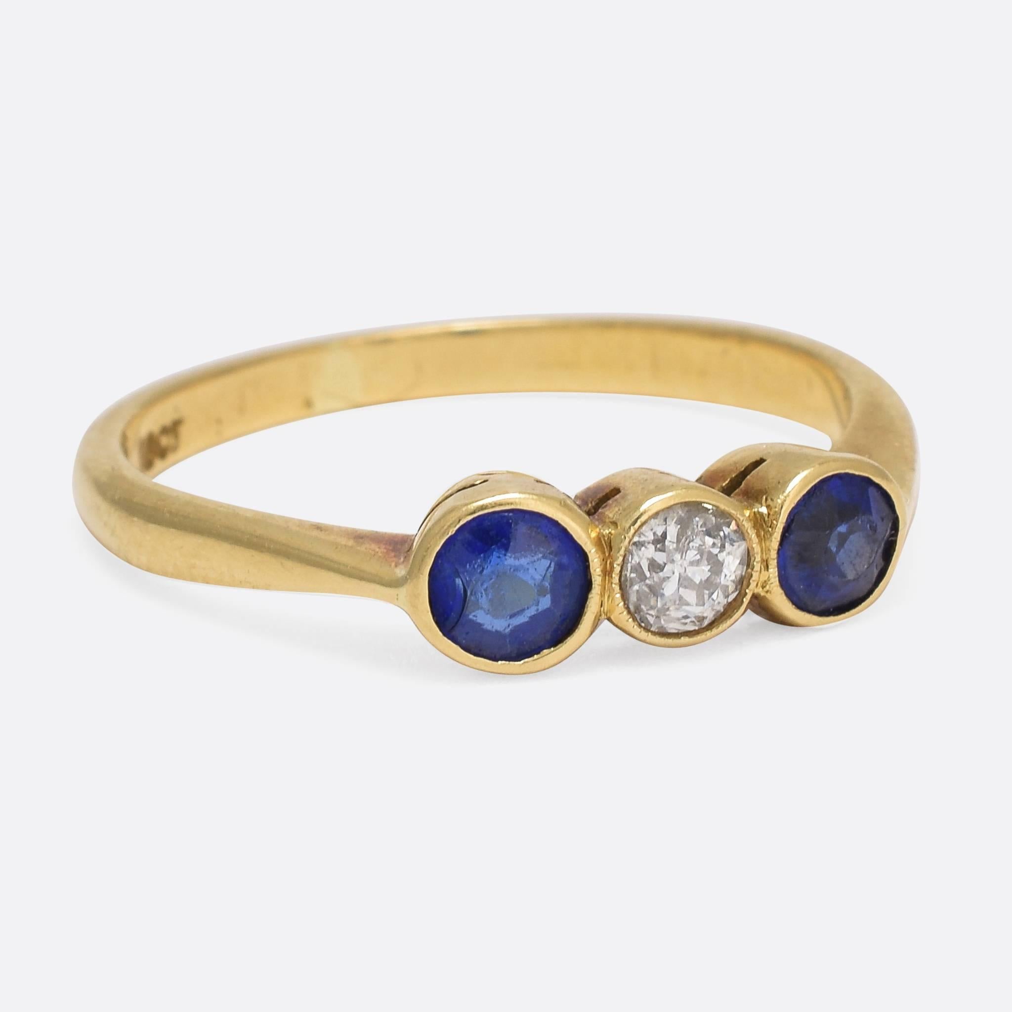 This pretty antique three-stone ring is set with a central old cut diamond, flanked by two vibrant blue sapphires (synthetic). The stones are bezel set, very typical of the period, with a subtle ring of millegrain around the diamond and cut-out